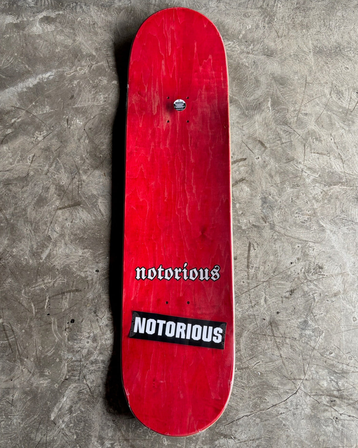 A red skateboard deck with 'notorious' branding in white text, displayed vertically against a textured gray concrete background.